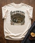 Please Assume I am Busy With a Very Long Book garment-dyed tee in Ivory, laid flat on dark oak beside a wool blanket, a beeswax candle and two pinecones