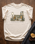 The Last Bus Was Two Hours Ago garment-dyed tee in Ivory, laid flat on dark oak beside a wool blanket, a beeswax candle and two pinecones