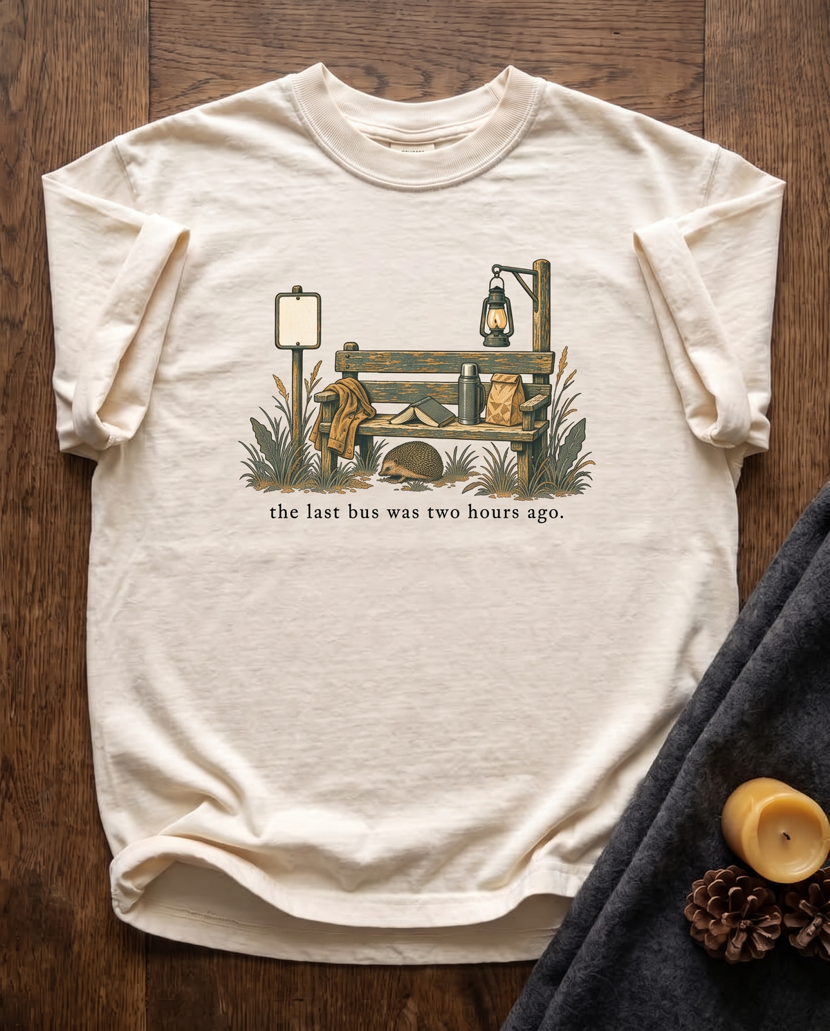 The Last Bus Was Two Hours Ago garment-dyed tee in Ivory, laid flat on dark oak beside a wool blanket, a beeswax candle and two pinecones