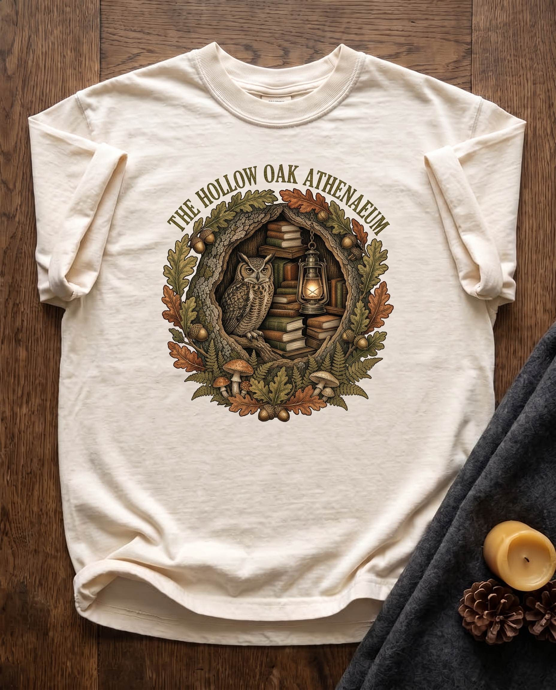 Hollow Oak Athenaeum garment-dyed tee in Ivory, laid flat on dark oak beside a wool blanket, a beeswax candle and two pinecones