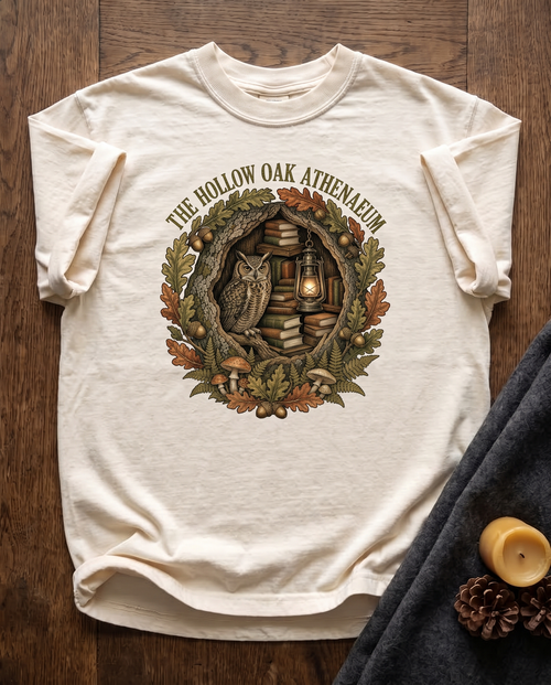 Hollow Oak Athenaeum garment-dyed tee in Ivory, laid flat on dark oak beside a wool blanket, a beeswax candle and two pinecones