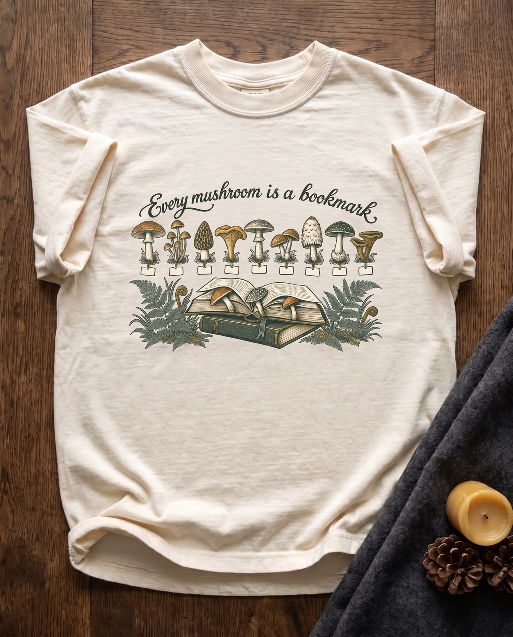 Every Mushroom Is a Bookmark garment-dyed tee in Ivory, laid flat on dark oak beside a wool blanket, a beeswax candle and two pinecones
