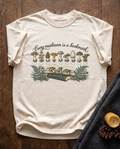 Every Mushroom Is a Bookmark garment-dyed tee in Ivory, laid flat on dark oak beside a wool blanket, a beeswax candle and two pinecones