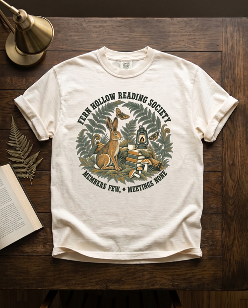 Fern Hollow Reading Society garment-dyed tee in Ivory, laid flat on dark oak beside an open book, a dried fern frond and a brass lamp