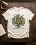 Membership Is One garment-dyed tee in Ivory, laid flat on dark oak beside an open book, a dried fern frond and a brass lamp