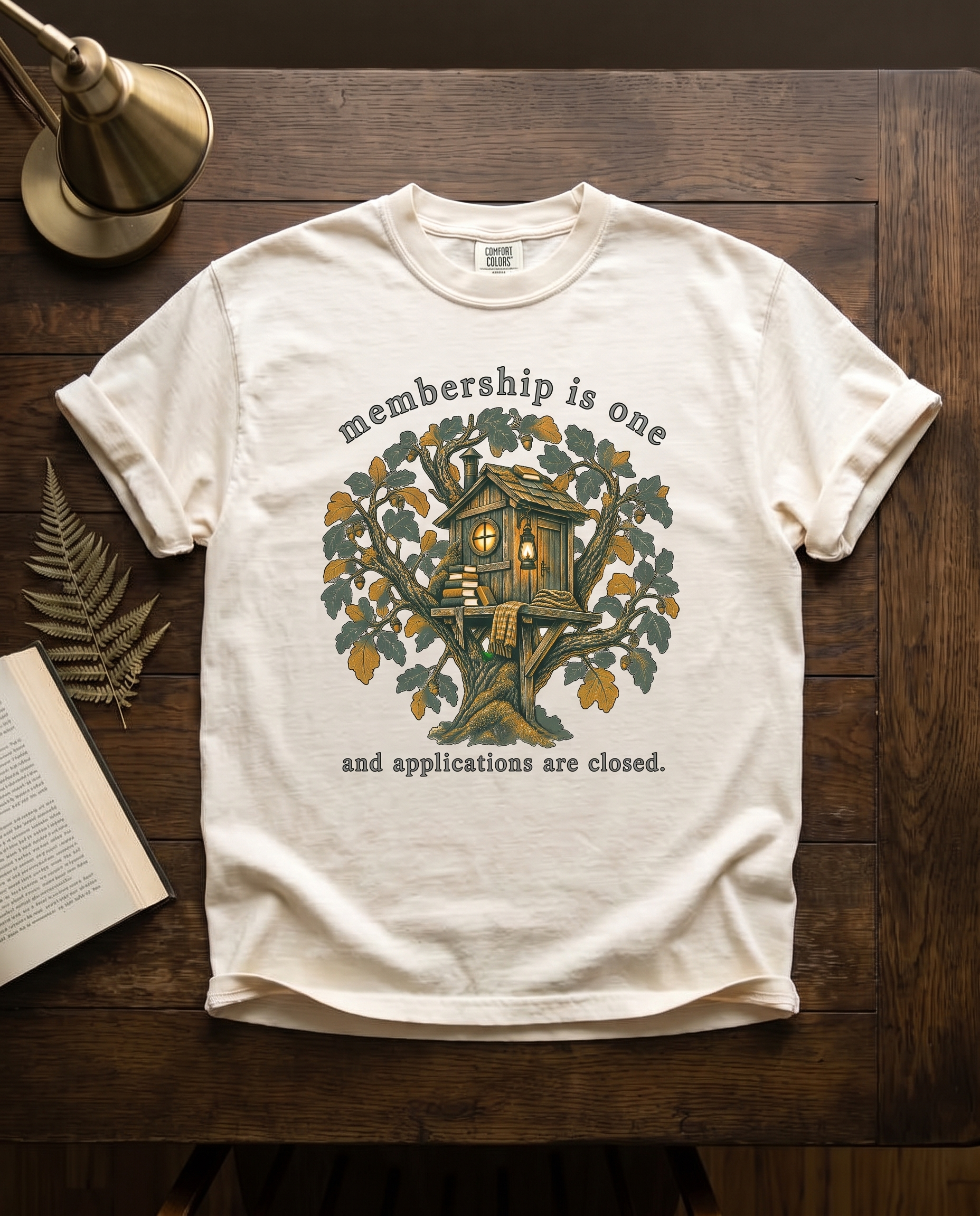 Membership Is One garment-dyed tee in Ivory, laid flat on dark oak beside an open book, a dried fern frond and a brass lamp