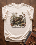 The Current Can Wait garment-dyed tee in Ivory, laid flat on dark oak beside cloth-bound books, reading glasses and scattered acorns