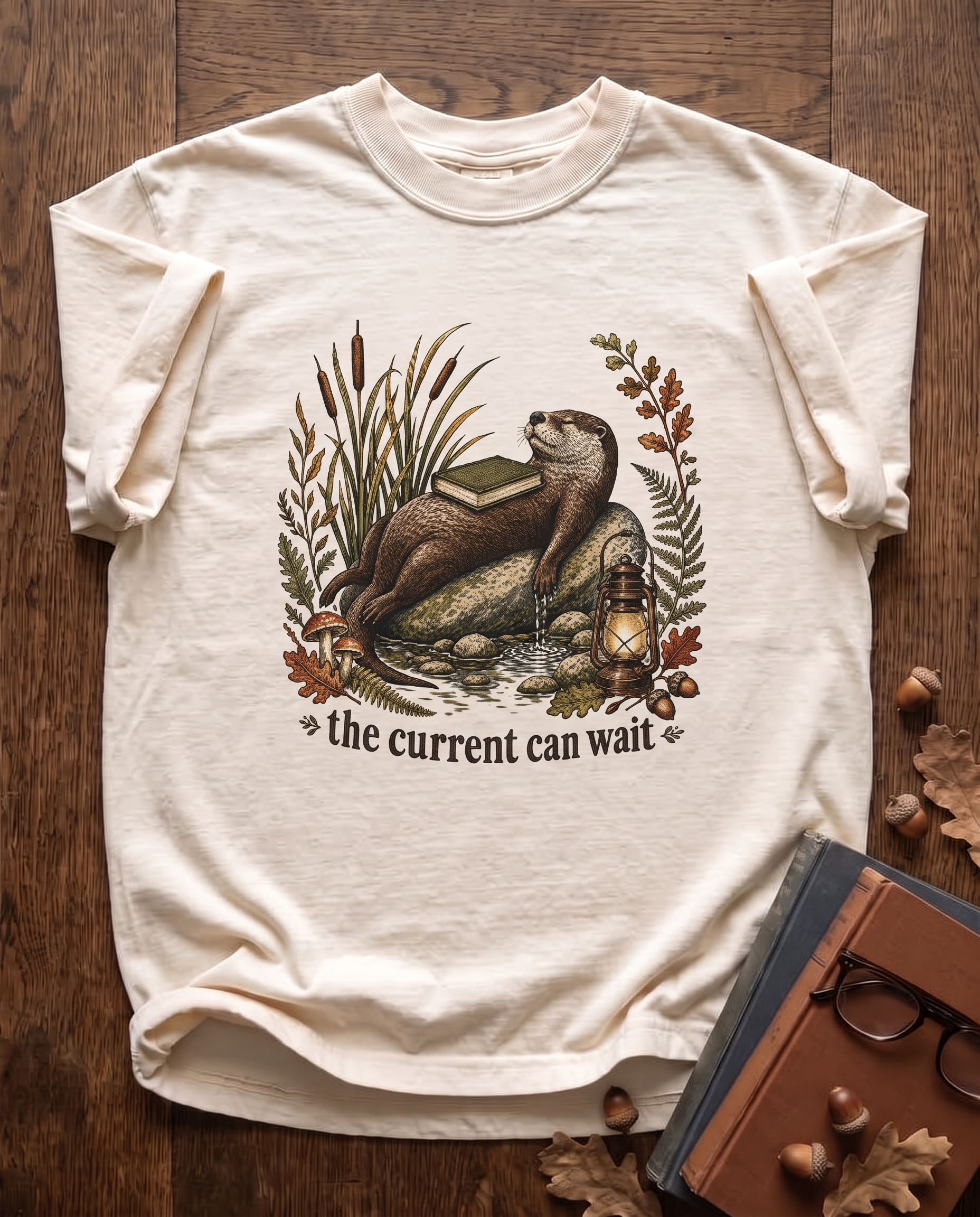 The Current Can Wait garment-dyed tee in Ivory, laid flat on dark oak beside cloth-bound books, reading glasses and scattered acorns