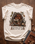 Hoarding Novels, Not Nuts garment-dyed tee in Ivory, laid flat on dark oak beside cloth-bound books, reading glasses and scattered acorns