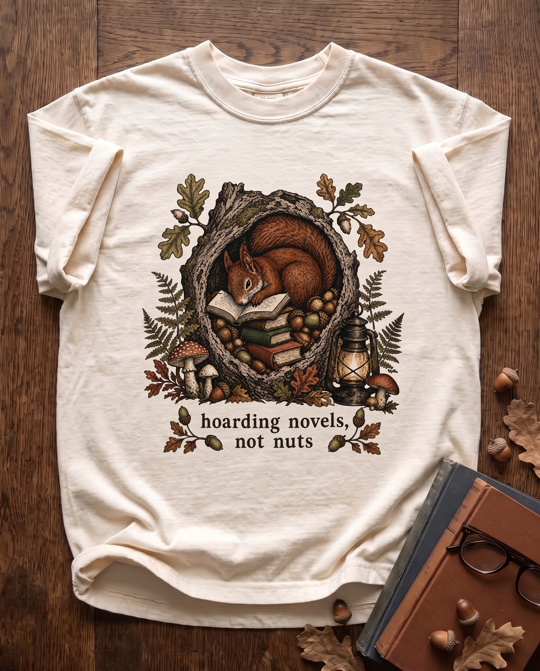 Hoarding Novels, Not Nuts garment-dyed tee in Ivory, laid flat on dark oak beside cloth-bound books, reading glasses and scattered acorns