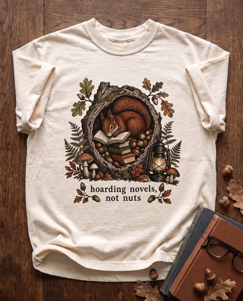 Hoarding Novels, Not Nuts garment-dyed tee in Ivory, laid flat on dark oak beside cloth-bound books, reading glasses and scattered acorns