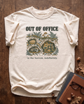Out of Office, In the Burrow garment-dyed tee in Ivory, laid flat on dark oak beside cloth-bound books, reading glasses and scattered acorns